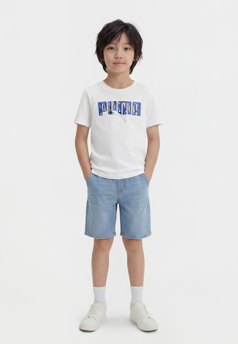 Αγόρι με μαύρα μαλλιά στέκεται με τα χέρια στις τσέπες, φορώντας λευκό μπλουζάκι με λογότυπο Puma, ανοιχτόχρωμο μπλε τζιν σορτς, λευκές κάλτσες και λευκά αθλητικά παπούτσια.