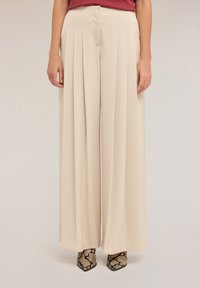 Pantalon large beige avec plis avant, tissu lisse, taille haute et coupe cintrée, associé à des chaussures texturées à imprimé serpent.