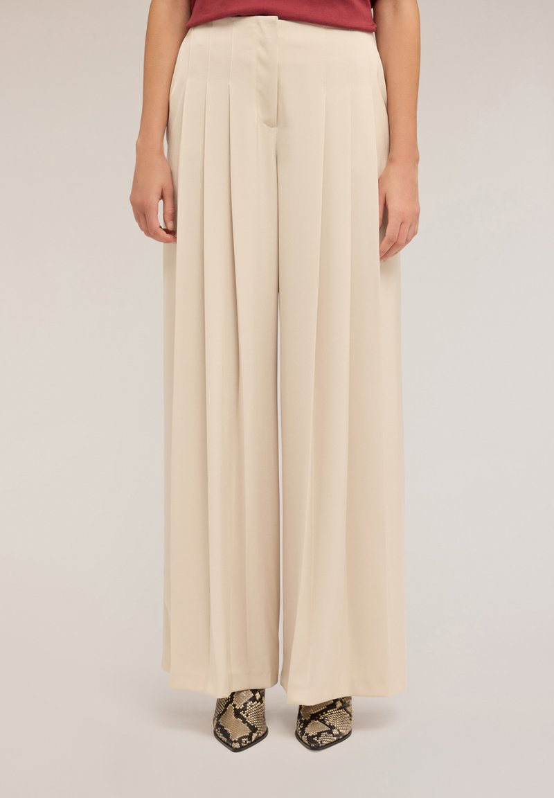 Pantalon large beige avec plis avant, tissu lisse, taille haute et coupe cintrée, associé à des chaussures texturées à imprimé serpent.