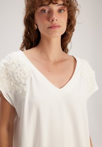 Weißes V-Ausschnitt-Oberteil mit floralen Applikationen an den Schultern, aus weichem Material und mit kurzen Ärmeln, für einen entspannten Sitz.