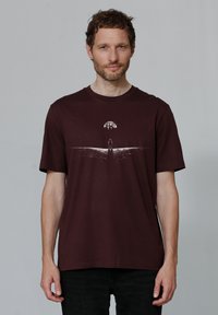 Bordeaux T-shirt, ronde hals, korte mouwen. Heeft een witte afbeelding van een figuur en een boom op een golvend ontwerp. Zachte katoenkwaliteit.