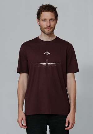 T-shirt bordeaux, col ras du cou, à manches courtes. Présente un graphisme blanc représentant une silhouette et un arbre sur un design ondulé. Matière en coton doux.