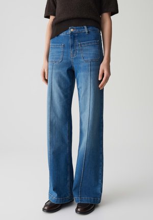 Brede, blauwe denim jeans met een hoge taille, voorzien van twee achterzakken en zichtbare stiksels, gecombineerd met donkere schoenen.