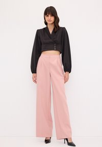 Schwarze, kurz geschnittene Bluse mit langen Puffärmeln und vorderen Knöpfen, kombiniert mit hoch taillierten, weit geschnittenen rosa Hosen und schwarzen spitzen Heels.