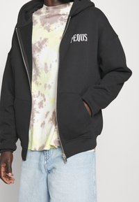 Henkilö, jolla on päällään musta vetoketjullinen huppari, jossa on "PEQUS"-logo, värjätty pastellinvärinen paita ja vaaleansiniset farkut, käsi hupparin taskussa.