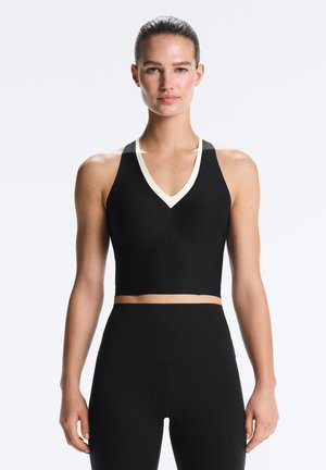 Femme aux cheveux tirés en arrière, portant un top de sport court noir avec un liseré blanc et des leggings noirs taille haute, face à l'avant.