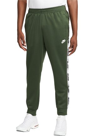 Mężczyzna ubrany w oliwkowo zielone joggery Nike z białym logo na udzie i wzorzystym paskiem po bokach, w połączeniu z białymi trampkami i białą koszulką.