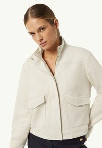 Kremowa kurtka z wysokim kołnierzem, przednim zamkiem błyskawicznym i dwoma dużymi kieszeniami. Gładka tekstura, usztywniony kształt i minimalistyczne detale designu.