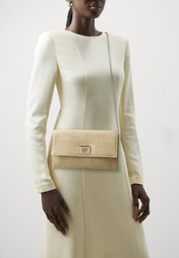 Beige geweven clutch tas met een omgevouwen klep en metalen sluiting. Voorzien van een zilveren schouderband en een gestructureerd oppervlak.
