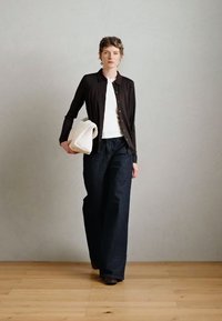 Mørk knap-front cardigan over en hvid top, parret med mørke denim jeans med brede ben. Holder en lysefarvet taske, står på et trægulv.