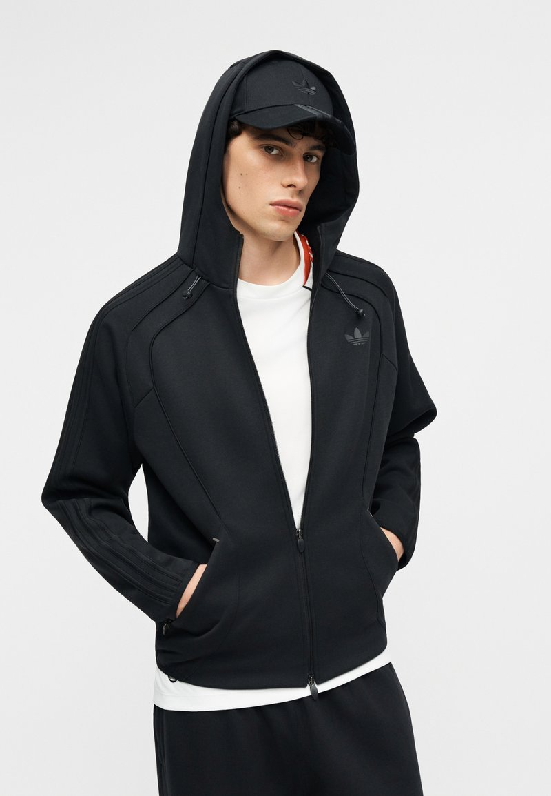 Jonge man met een zwart vest met rits en capuchon omhoog, zwarte pet, wit T-shirt en zwarte broek, staand met handen in de zakken tegen een witte achtergrond.