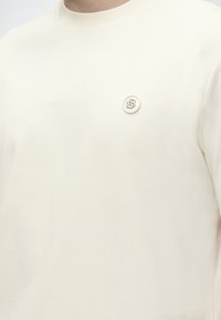 Μπλούζα σε κρεμ χρώμα με στρογγυλή λαιμόκοψη και μικρό στρογγυλό σήμα με τα γράμματα "SB" στο αριστερό στήθος.