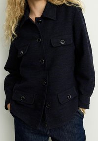 Mørkeblå, tekstureret jakke lavet af vævet stof, med en krave, forlommer og sorte knapper. Casual fit design.