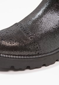 Primer plano de la parte delantera de una bota negra brillante con tela texturizada y una suela de goma negra gruesa con profundos relieves.
