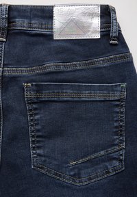 Jeans de mezclilla azul oscuro con una etiqueta plateada que dice "SE UN ESPEJO", costuras visibles y un único bolsillo trasero con una pequeña marca de desgaste.