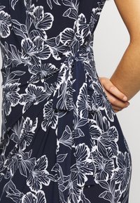 Marinblå klänning med vitt blommönster, knuten med en rosett i midjan, med en hand vilande på höften.