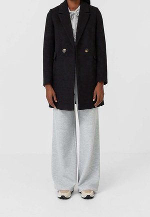 Manteau croisé en mélange de laine noire avec de gros boutons, associé à un pantalon de survêtement gris clair à jambes larges et des baskets beiges.