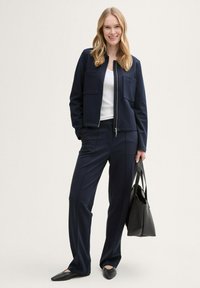 Mörkblå zip-up jacka med två framfickor, i kombination med matchande skräddarsydda byxor. Modellen bär en svart tote-väska och har på sig svarta flats.
