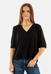 Top nero a V con maniche corte, realizzato in un tessuto testurizzato. Abbinato a jeans blu, con una leggera lucentezza visibile nel materiale.