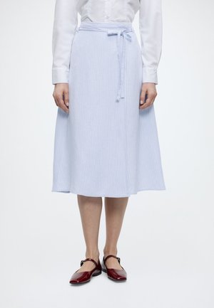 Frau trägt einen hellblauen, knöchellangen, gestreiften Rock mit gebundenem Taillenband und burgunderrote Mary-Jane-Schuhe, steht vor einem schlichten weißen Hintergrund.