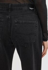 Čierne rifľové nohavice z denimovej tkaniny so hladkým povrchom, klasickým päťvreckovým dizajnom a pásom s logom. Pohľad zozadu ukazuje šikmo umiestnené zadné vrecká.