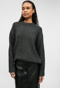 Γκρι πλεκτό πουλόβερ με υφή ριμπ, στρογγυλή λαιμόκοψη και άνετη εφαρμογή. Φοριέται πάνω από μια μαύρη δερμάτινη φούστα.