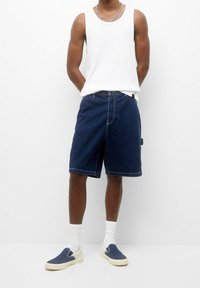 Shorts de mezclilla en azul oscuro, con un corte holgado y costuras en contraste. Combinados con una blusa sin mangas blanca y zapatos azul claro tipo slip-on.