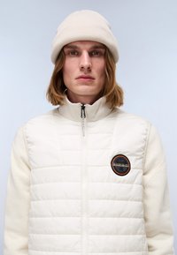 Weißer gesteppter Jacket mit hohem Kragen, der einen gerippten cremefarbenen Pulloverbereich und ein rundes Logo-Patch auf der Brust aufweist. Ergänzt wird das Outfit durch eine beigefarbene Mütze.