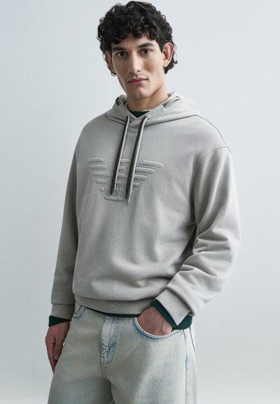 Sweat à capuche gris clair avec un logo embossé, cordons ajustables et poignets côtelés. Associé à un jean en denim bleu clair.