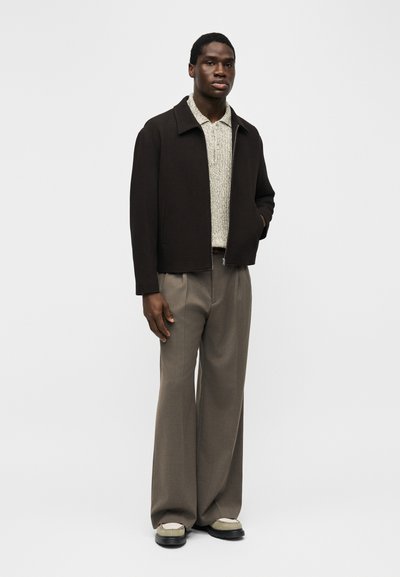 Νεαρός άνδρας φοράει μαύρο μπουφάν με φερμουάρ πάνω από μπεζ πλεκτό πόλο, καφέ παντελόνια με φαρδιά πόδια και μπεζ και μαύρα παπούτσια, στέκεται μπροστά σε μονόχρωμο φόντο.