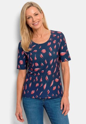 Blonde Frau lächelt, trägt ein marineblaues Kurzarmoberteil mit rotem Pinselstrichmuster und blaue Jeans, steht vor hellem Hintergrund.