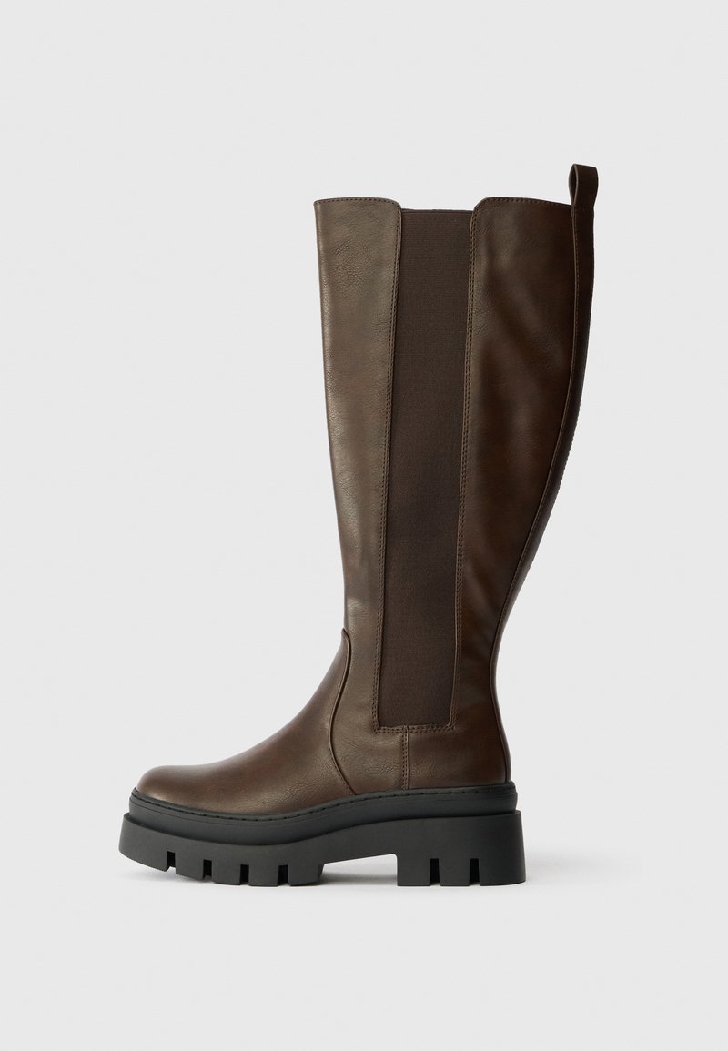 Brown leather knee-high boot with a matte finish, elastic side panels, and a chunky black rubber sole. Straight shape with a rounded toe.