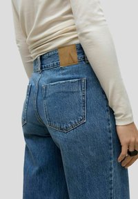 Kõrgvöödiga sinised denim teksad, millel on kulunud pesu, kaks tagataskut, nahkne silt vööl ja sirge lõige.