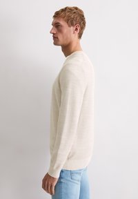 Jasnobeżowy sweter z okrągłym dekoltem o fakturowanym wzorze dzianiny. Długie rękawy i prążkowane mankiety. Noszony z jasnoniebieskimi dżinsami.