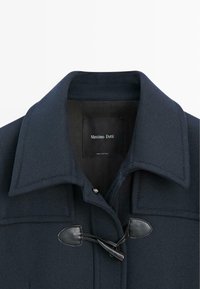 Cappotto di lana blu navy con colletto a punta, chiusura a toggle con dettagli in pelle e fodera interna liscia. Etichetta del marchio visibile.
