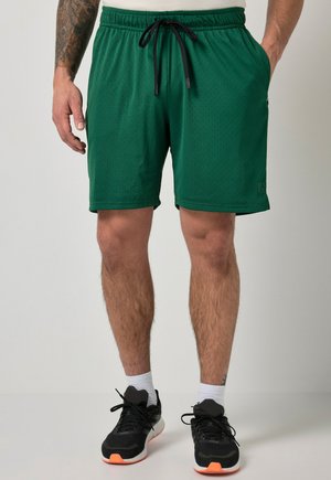 Mand iført grønne atletikshorts med sort snøre, hvide strømper og sorte løbesko, stående mod en ensfarvet lys baggrund.