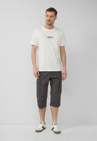 T-shirt en coton blanc avec logo navy, associé à un short cargo gris foncé jusqu'aux genoux avec poches latérales, et des baskets blanches avec des accents gris.