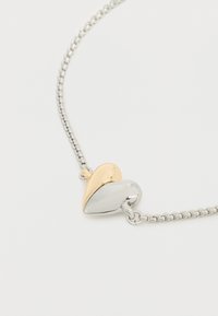 Μεταλλικό μενταγιόν διπλής καρδιάς με το ένα μισό χρυσό και το άλλο μισό ασημένιο, σε ασημένια αλυσίδα κολιέ σε απλό φόντο.