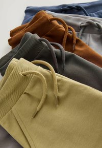 Fünf Paar Jogginghosen in verschiedenen Farben: Gelb, olivgrün, grau, rostfarben und marineblau. Mit Kordelzügen und gerippten Taillenbändern. Weiches Material.