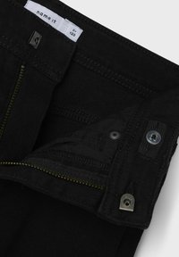 Pantalones de mezclilla negros con cierre de cremallera, cremallera de acento dorado y cierre de botón. Forro interior visible con detalles de costura.