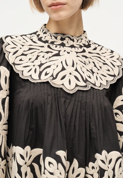 Femme portant une robe noire avec une broderie florale crème complexe sur le col montant, la poitrine, les manches et l'ourlet.