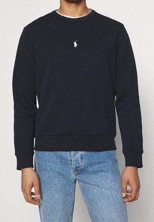 Mies pukeutunut mustaan pitkähihamaiseen collegepaitaansa, jossa on pieni valkoinen poloilijan logo rinnassa keskellä, ja vaaleansinisiin farkkuihin.