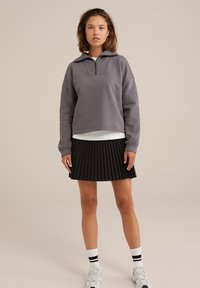 Grå bomulds sweatshirt med lynlås ved kraven, parret med en sort plisseret nederdel. Modellen bærer hvide strømper med sorte striber og sporty sko.