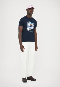 Tmavě modré bavlněné tričko s grafickou potiskem, spárované s bílými kalhotami a černými botami. Světle růžová čepice doplňuje outfit.