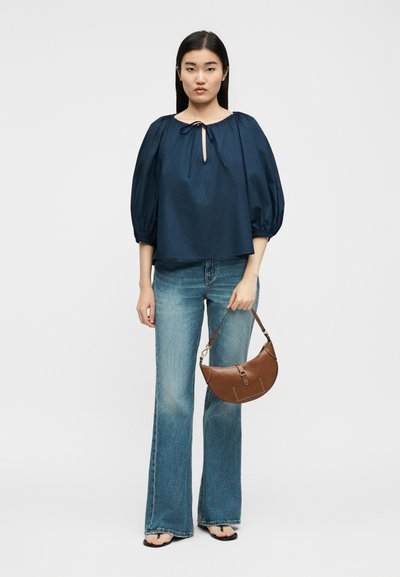 Žena v tmavomodrej blúzke s nafúknutými rukávmi, modré rozšírené džínsy, čierne žabky, nesie hnedú koženú polmesiacovú kabelku.