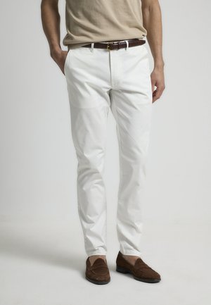 Pantalones de algodón blancos con un corte slim, que cuentan con un cierre de cremallera y bolsillos frontales. Combinados con unos mocasines de ante marrón y un cinturón marrón.