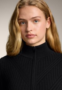 Plavuša plavih očiju u crnom rebrastom džemperu na patentni zatvarač, gleda ravno ispred sebe s neutralnim izrazom na licu, na jednobojnoj pozadini.