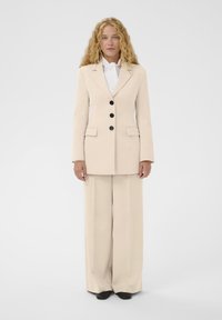 Costume beige clair sur mesure comprenant un blazer avec des boutons noirs, des revers crantés et un pantalon à jambes larges. Texture de tissu lisse, design minimaliste.