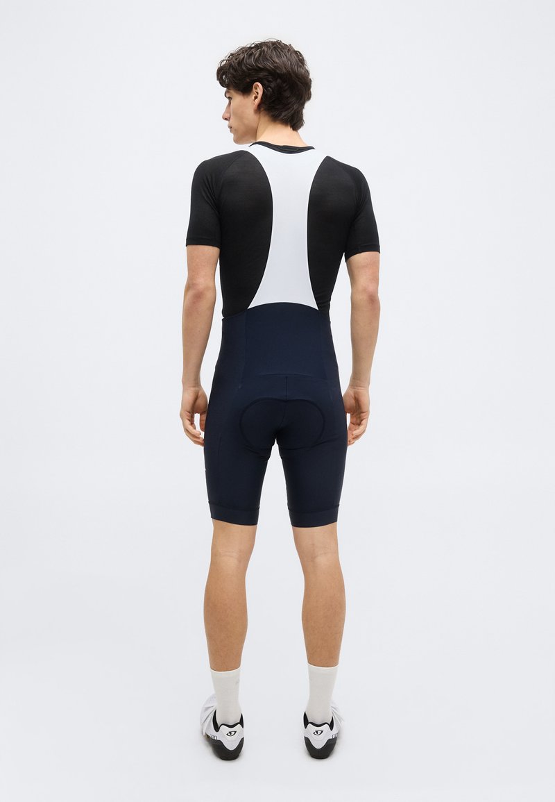 Man wearing black and white short-sleeve cycling bib shorts and white socks, standing facing away against a plain background.