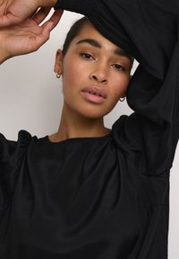 Black, long-sleeve blouse made of smooth fabric, featuring puffed sleeves and a rounded neckline. Model poses with one arm raised.